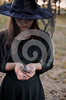 Young cute beautiful woman in dark dress and witch`s hat holdsblack spider in her hands. Halloween party costume. Forest