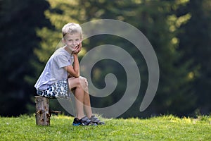 Young cute blond child boy sitting on tree stump on green grassy clearing on bright summer day