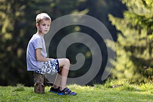 Young cute blond child boy sitting on tree stump on green grassy clearing on bright summer day