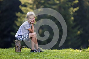 Young cute blond child boy sitting on tree stump on green grassy clearing on bright summer day