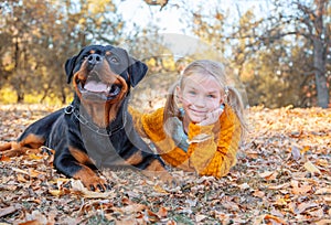 Young cute blonde girl child and her German Rottweiler dog lying and posing in autumn leaves. Friendship, pet and kid.