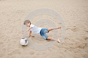 Young cute boy in denim shorts and a white T-shirt, jumps for a soccer ball on the sand