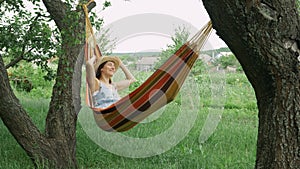 Young cute brunette girl resting on hammock, unwinds on hammock in countryside. Caucasian woman in hat and dress relaxing and rais