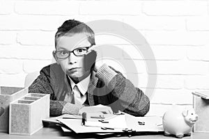 Young cute businessboy in grey sweater and glasses sitting at table with papers pink piggy pig bank holding and speaking