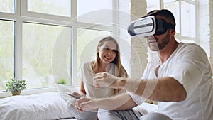 Young cute couple with tablet computer and virtual reality headset playing 360 VR video game while sitting in bed at