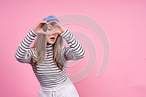 Young cute emotional caucasian lady in a fashionable beret shows heart on a pink background