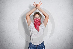 Young cute female feminist wears red bandana and casual grey t shirt, protects womens rights, keeps hands raised and crossed, look