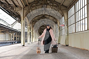 Young cute girl with dog and suitcases on platform waiting for train in pink
