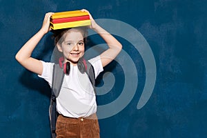 young cute  girl with headphones  with pile of books on head on blue wall background