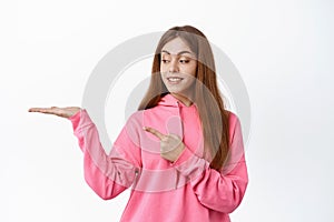 Young cute girl holding your logo on hand, pointing and looking at palm with empty space copyspace, standing against
