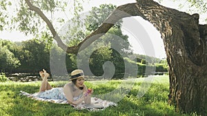 Young cute girl relaxing on blanket on grass under big tree and blowing bubbles at sunny summer day. Charming female in hat and dr
