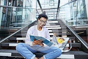 Young cute indian girl at university building sitting on stairs reading a book, wearing hipster glasses, lifestyle