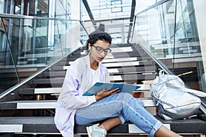 Young cute indian girl at university building sitting on stairs reading a book, wearing hipster glasses, lifestyle