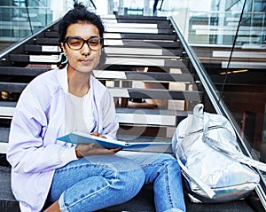 Young cute indian girl at university building sitting on stairs reading a book, wearing hipster glasses, lifestyle