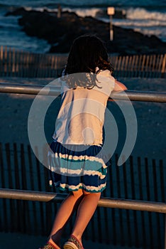 Young cute little girl on the boardwalk with back to camera looking towards the ocean surf