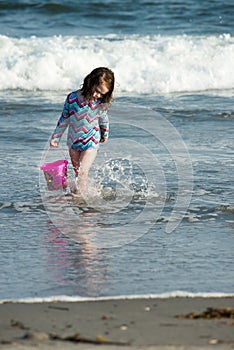 Young cute little girl playing at the seaside carrying a red bucket at the edge of the surf on a sandy beach in summer