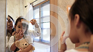 Young cute little girl standing on chair in the bedroom in morning and smile looking at mirror while mother combing her hair.