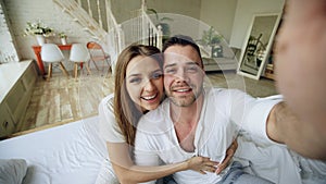 Young cute and loving couple having video chat holding tablet computer and chatting to parents sitting in bed at home