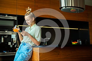A young cute modern girl with smartphone and a glass of juice is relaxing in a cozy kitchen