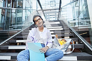 Young cute modern indian girl at university building sitting on stairs reading a book, wearing hipster glasses