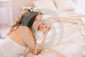 Young cute mom in a wreath of cherry blossoms in a white dress holding a baby in her arms in a spring bedroom