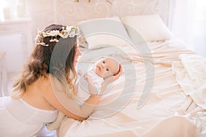 Young cute mom in a wreath of cherry blossoms in a white dress holding a baby in her arms in a spring bedroom