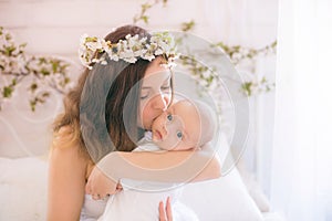 Young cute mom in a wreath of cherry blossoms in a white dress holding a baby in her arms in a spring bedroom