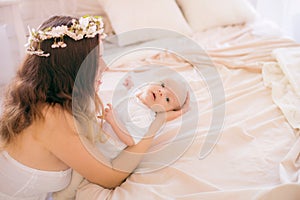 Young cute mom in a wreath of cherry blossoms in a white dress holding a baby in her arms in a spring bedroom