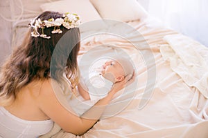 Young cute mom in a wreath of cherry blossoms in a white dress holding a baby in her arms in a spring bedroom