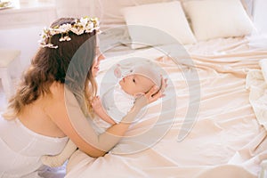 Young cute mom in a wreath of cherry blossoms in a white dress holding a baby in her arms in a spring bedroom