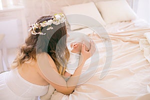 Young cute mom in a wreath of cherry blossoms in a white dress holding a baby in her arms in a spring bedroom