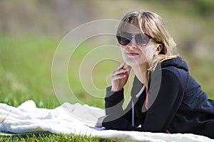 Young cute pretty blond girl in black sweater and dark sunglasses lies on white towel on green grass enjoying nature and warm sun