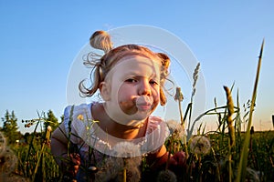 Young cute pretty funny girl in a meadow with dandelion flowers in the light of the sun at sunset and blue sky background. Girl