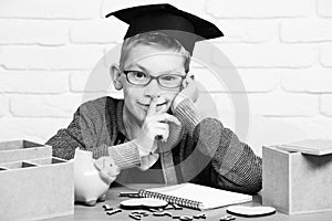 Young cute pupil boy in grey sweater and glasses sitting at desk with copybook wooden numbers pink piggy pig bank in