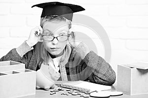 Young cute pupil boy in grey sweater and glasses sitting at desk with copybook wooden numbers pink piggy pig bank in