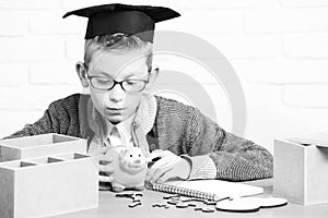 Young cute pupil boy in grey sweater and glasses sitting at desk with copybook wooden numbers pink piggy pig bank in