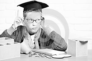 Young cute pupil boy in grey sweater and glasses sitting at desk with copybook wooden numbers pink piggy pig bank in