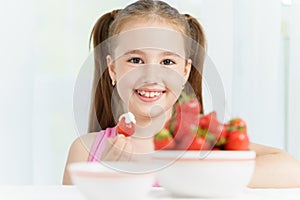 Young cute smiling european little girl is eating ripe jucy strawberry with sour cream and holding white plate of many