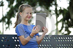 Young cute smiling teenager girl looking at screen of digital tablet sitting on bench in park on summer sunny day