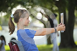 Young cute smiling teenager girl looking at screen of digital tablet sitting on bench in park on summer sunny day