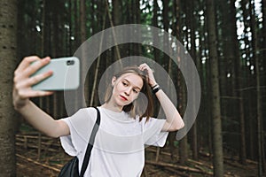 Young cute tourist girl in the mountain forest using smartphone to take a selfie. Cheerful attractive female traveler taking