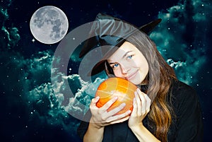 A young cute witch on Halloween with a pumpkin on the background of a creepy dark magical sky. A girl in a witch`s hat hugs a pump