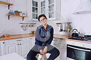 Young cute woman 30+ with short hair in blue jeans and a shirt sitting and relaxing in her clean kitchen
