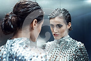 Young cyber woman in silver futuristic costume looking in the mirror.