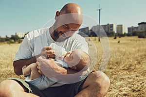 Young dad gives to eat a newborn baby from a bottle with a dummy sitting on the grass on the nature. happy father is wearing