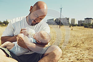 Young dad gives to eat a newborn baby from a bottle with a dummy sitting on the grass on the nature. happy father is wearing