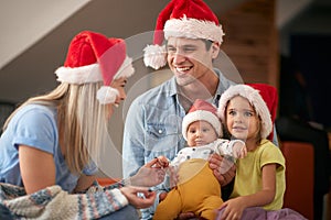 Young dad holding in lap daughter and a baby, smiling, looking at blonde mom, wearing santa hats. christmastime
