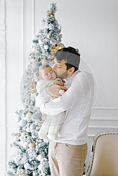 A young dad holds a baby in his arms and kisses it near the Christmas tree in the living room in the house