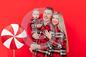 Young dad, mom and little son in red plaid pyjamas on red Christmas background with big lollipop. Amazing happy family