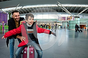 Young dad plays with his son, rolls him on suitcase in departure hall of international airport, waiting for passport and customs